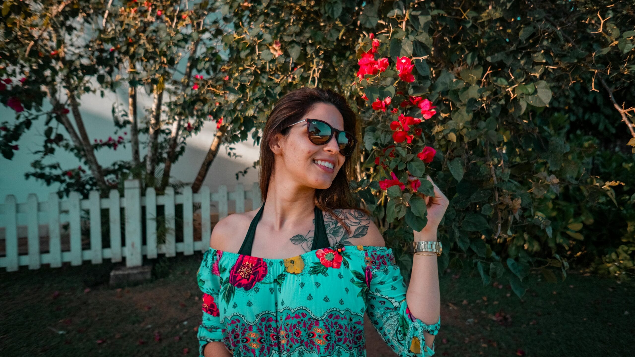 Smiling woman in vibrant floral dress enjoying a sunny day among red flowers in the garden.